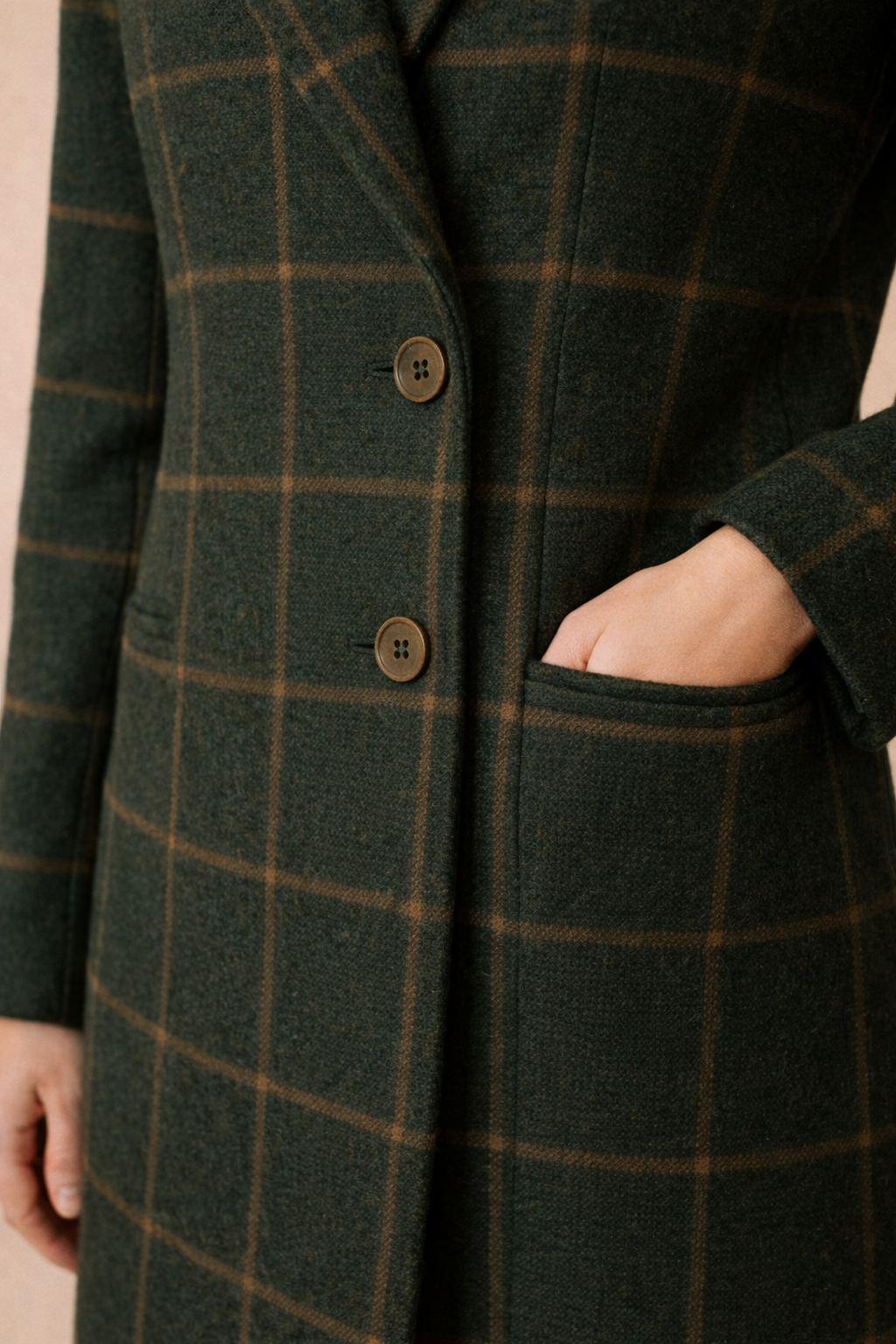 Detalle de los dos botones del abrigo Cashia Copenhague verde oscuro de cuadros, colocados en línea vertical en el cierre frontal, con bolsillo lateral visible sobre fondo nude.