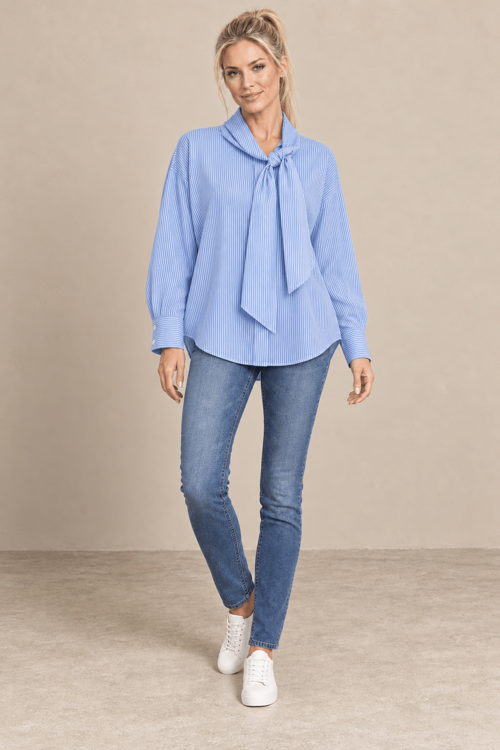 Camisa Cashia Elisa azul de raya fina blanca y cuello con lazada, vista frontal de cuerpo entero con vaqueros.
