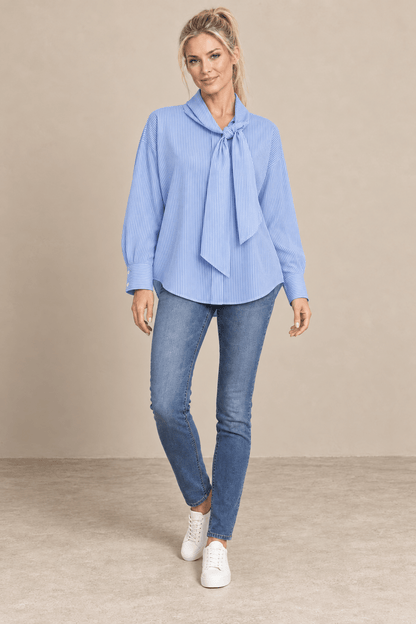 Camisa Cashia Elisa azul de raya fina blanca y cuello con lazada, vista frontal de cuerpo entero con vaqueros.
