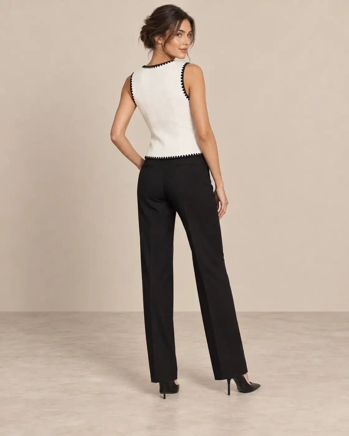 Chaleco Cashia Elegance blanco con ribete negro en talla S, sobre modelo Cashia morena con pelo semirecogido, pantalón de vestir negro y tacones negros, vista de espalda de cuerpo entero.