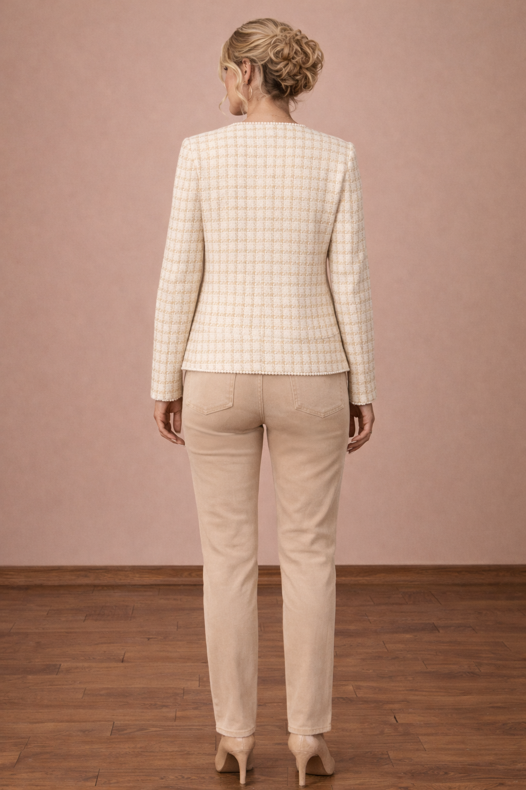 Chaqueta Cashia Capri en tweed beige con patrón de cuadros y ribete perlado, vista trasera cuerpo entero con corte recto, modelo rubia elegante con cabello recogido sobre fondo nude y suelo parquet marrón oscuro.