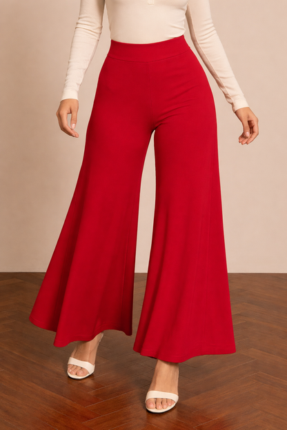 Pantalón Cashia Granada rojo en medio plano inferior, de cintura a talón, con pierna ancha y caída fluida sobre fondo nude y suelo de madera en espiga.