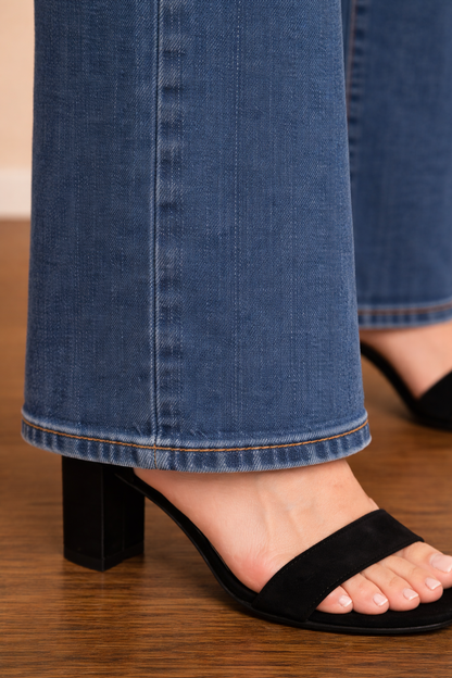 Detalle del bajo del Pantalón Cashia Málaga azul denim con corte acampanado y costura visible en el dobladillo, sobre suelo de parquet marrón oscuro.