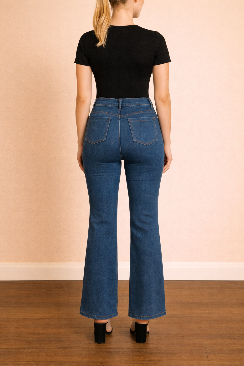 Pantalón Cashia Málaga azul denim de tiro alto y corte acampanado en vista trasera de cuerpo entero, con bolsillos posteriores y caída estructurada, sobre fondo nude con suelo de parquet marrón oscuro.