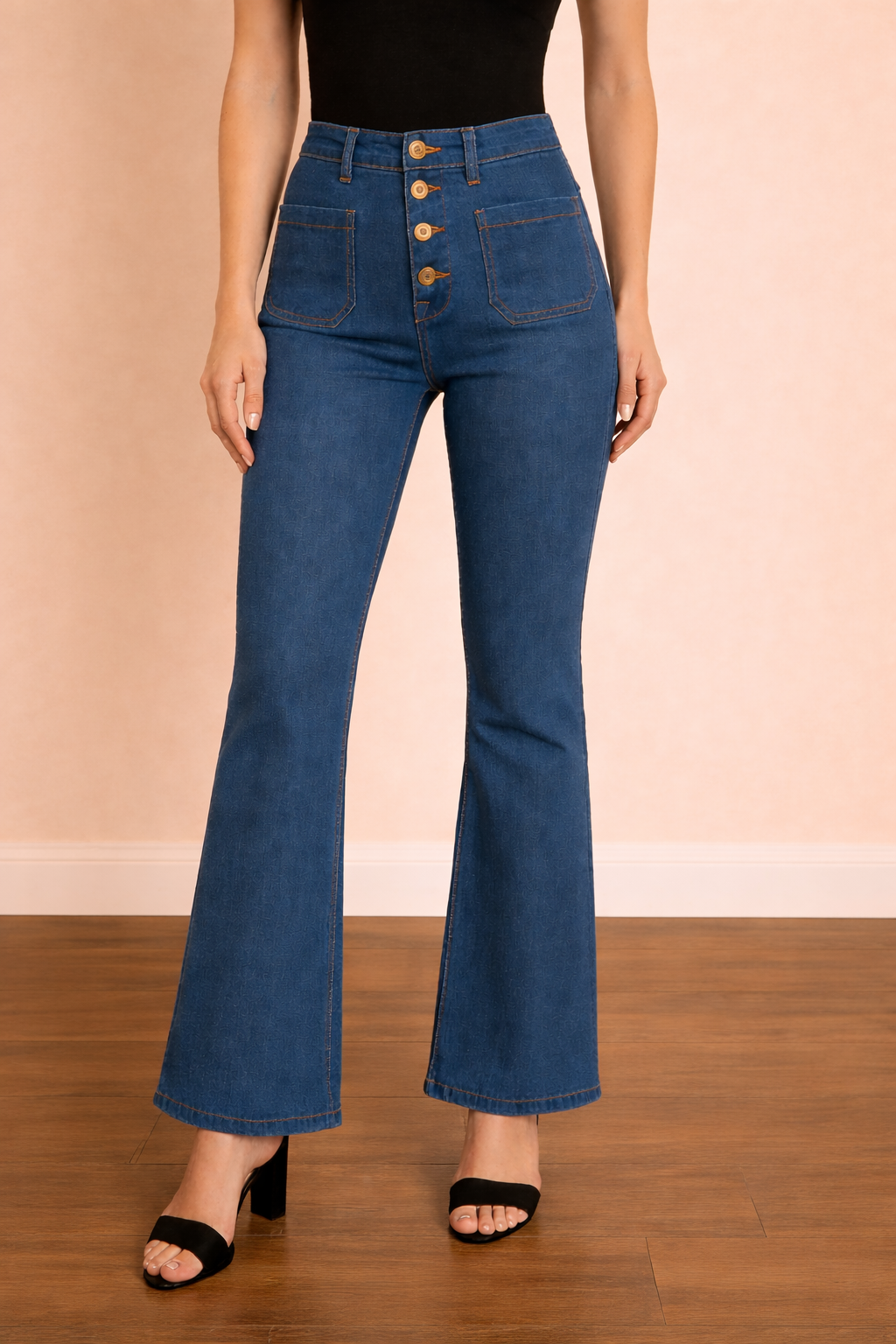 Pantalón Cashia Málaga azul denim en plano medio inferior, de cintura a tacón, con tiro alto, cierre frontal de botones visibles y corte acampanado, sobre fondo nude con suelo de parquet marrón oscuro.