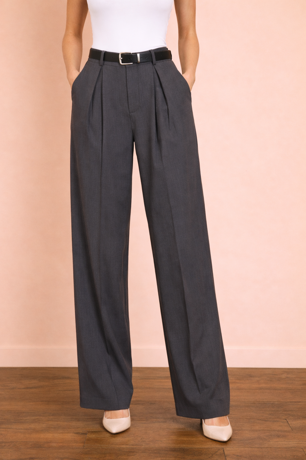 Pantalón Cashia Sevilla gris en plano medio inferior, de cintura a tacón, con tiro alto, pinzas delanteras y pernera ancha de caída fluida, sobre fondo nude con suelo de parquet marrón oscuro.