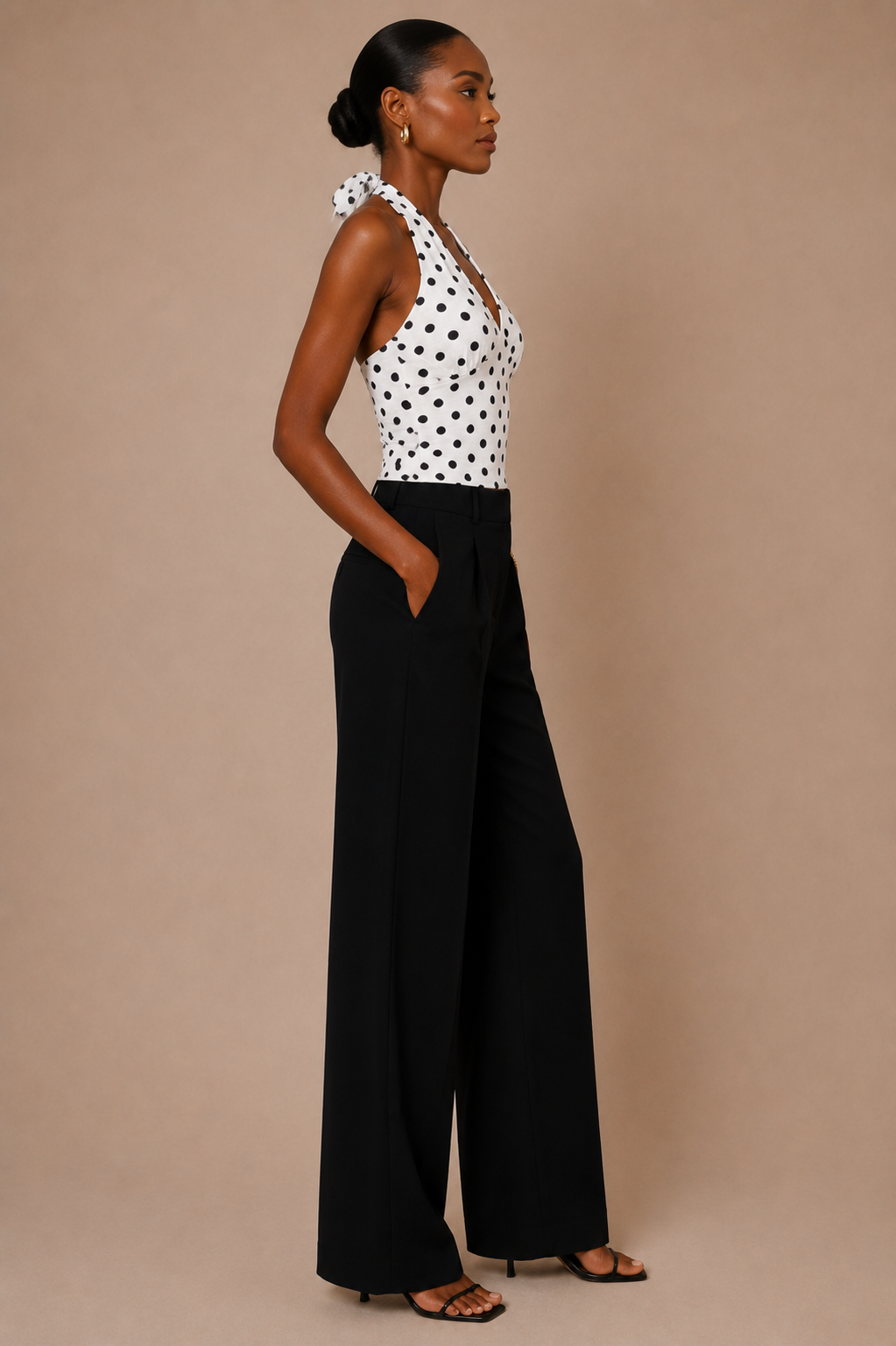 Vista de perfil del Top Cashia Amalfi blanco de lunares, con cuello halter anudado, combinado con pantalón negro sobre fondo nude.