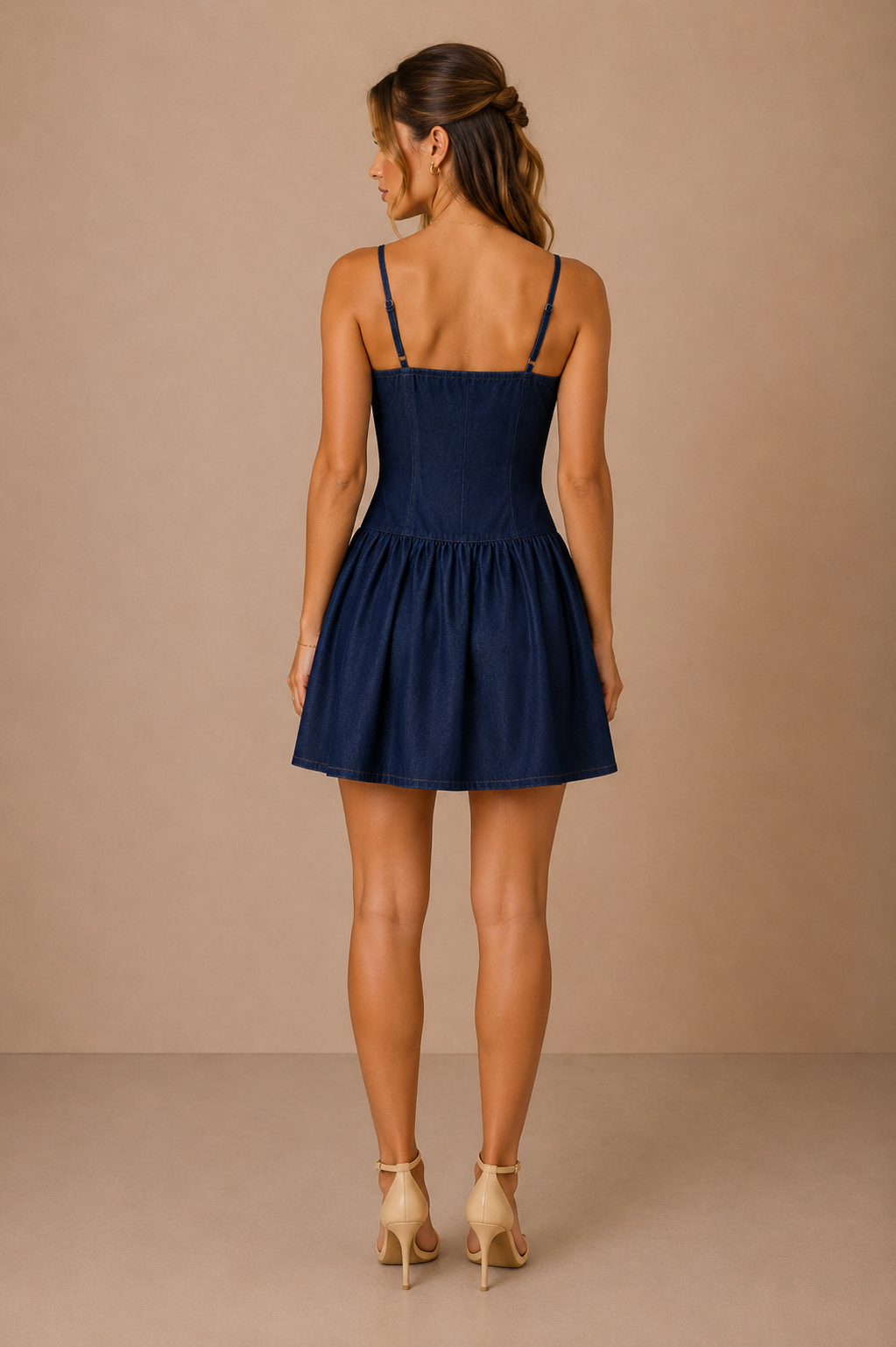 Modelo Cashia morena, elegante y maquillada, con pelo semirecogido. Lleva el Vestido Cashia Denim Málaga azul oscuro. Vista trasera de cuerpo entero mostrando la espalda y los tirantes finos sobre fondo nude con suelo definido.