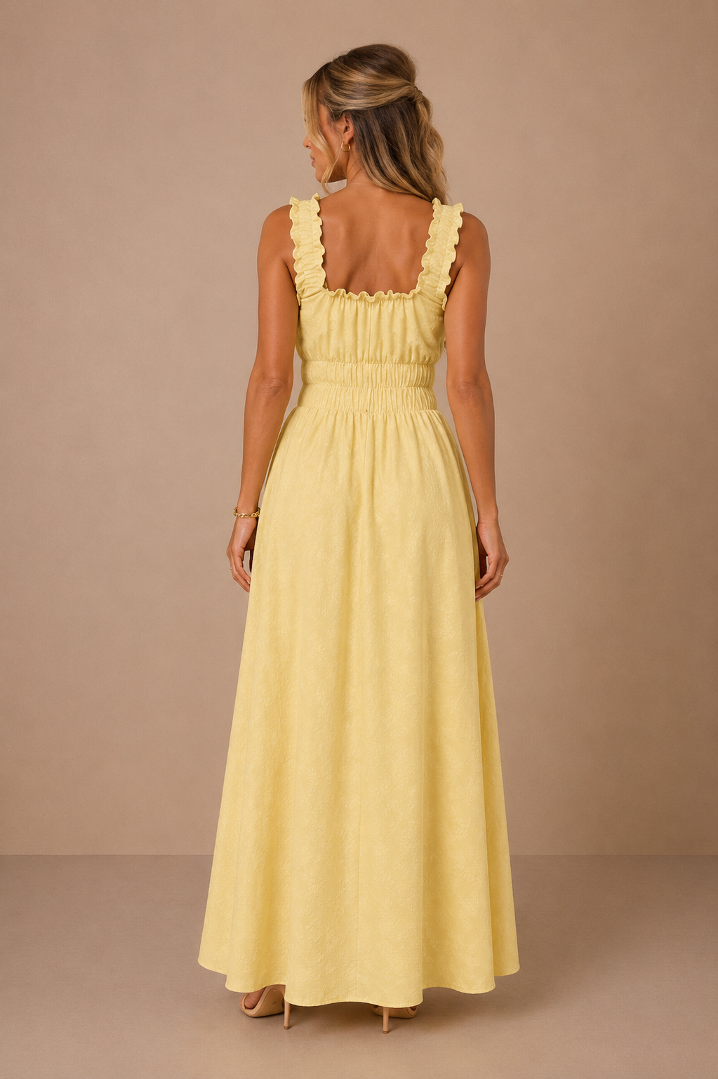 Modelo Cashia rubia de 40 años, elegante y maquillada, con pelo semirecogido. Lleva el Vestido Cashia Palermo amarillo. Vista trasera de cuerpo entero mostrando la espalda y los tirantes fruncidos sobre fondo nude con suelo definido.