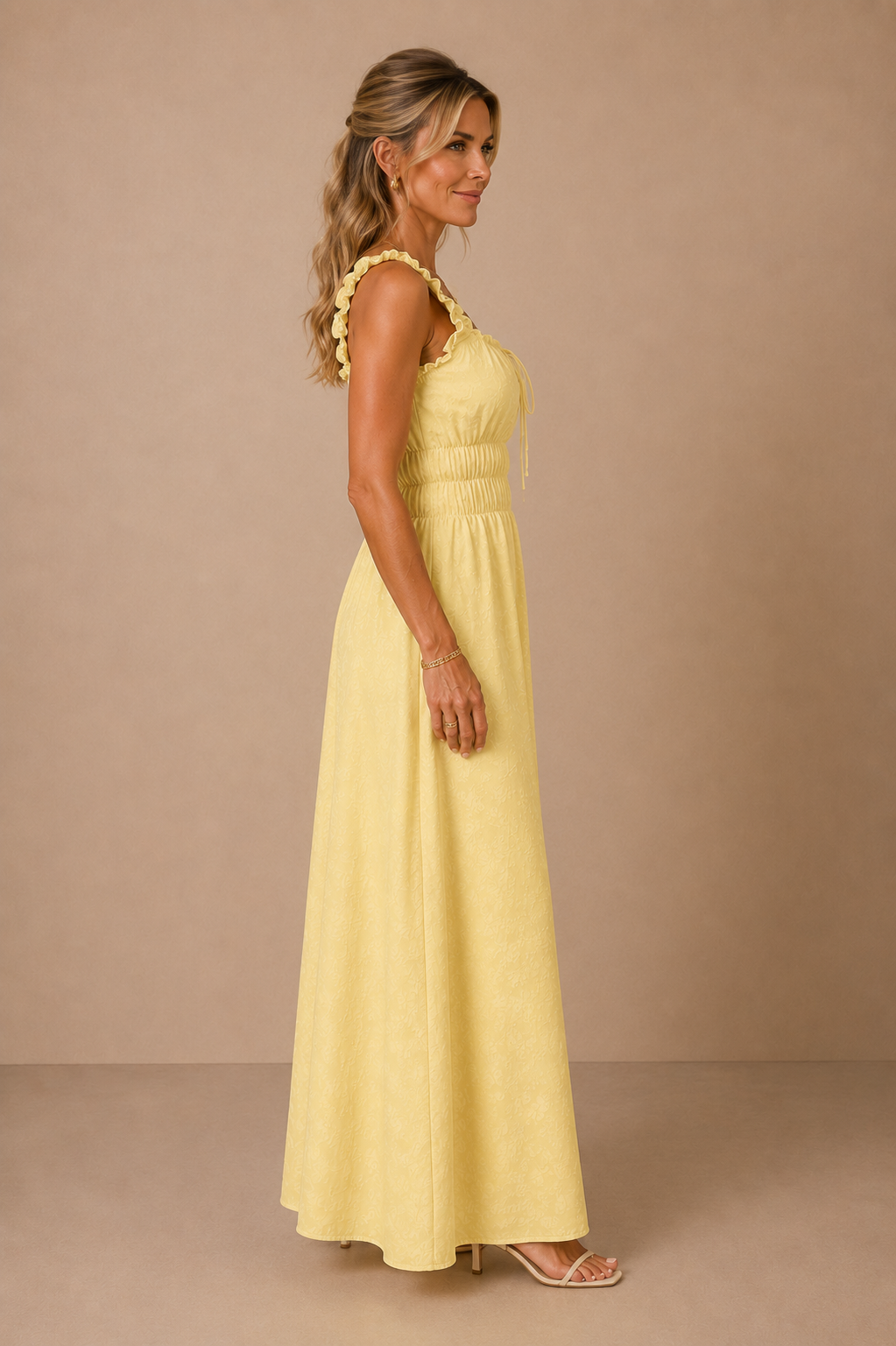 Modelo Cashia rubia de 40 años, elegante y maquillada, con pelo semirecogido. Lleva el Vestido Cashia Palermo amarillo. Vista lateral de cuerpo entero sobre fondo nude con suelo definido.